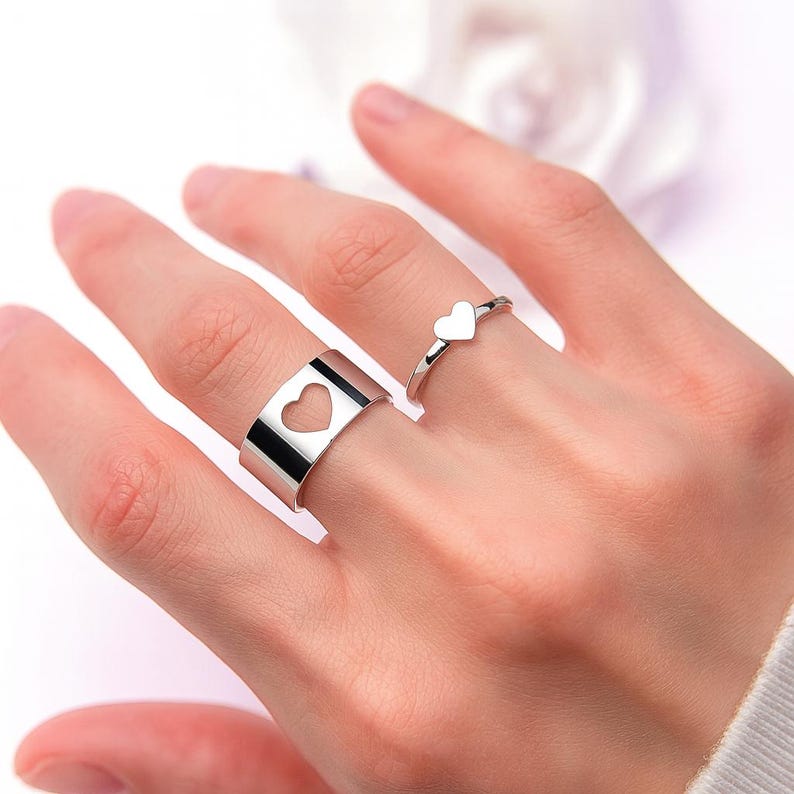 Op de afbeelding: Twee zilveren ringen worden op een hand getoond. De ene is een band met een hartvormige uitsparing. De andere is een dunne band met een klein hartje. De ringen zijn waarschijnlijk gemaakt van sterling zilver of een vergelijkbaar metaal.