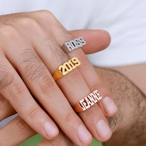 May include: Three rings with raised lettering spelling out "ROSS", "2019", and "JEANNE". The rings are gold and silver in colour. The rings are worn on the fingers.