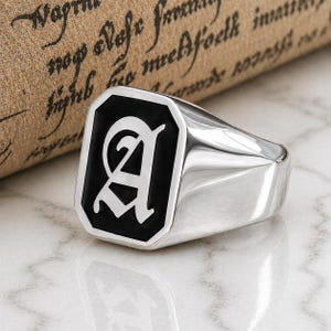 Puede incluir: Un anillo de sello plateado con una cara de esmalte negro que presenta la letra "A" en una fuente estilizada. El anillo tiene un acabado pulido y una cara rectangular. El fondo incluye un pergamino con texto descolorido.