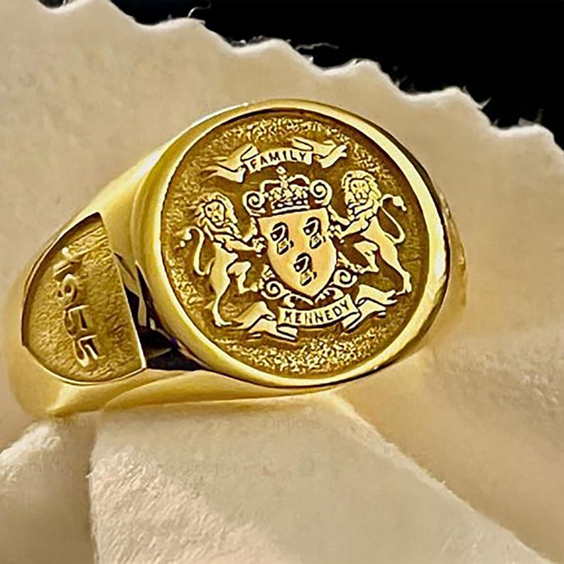 May include: A gold signet ring featuring a circular face with an engraved coat of arms, two lions, a crown, and the text FAMILY KENNEDY. The side of the ring band is engraved with the year 1955 in a bold, recessed font style.