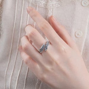 Puede incluir: Un anillo plateado con un dise&ntilde;o de plumas. El anillo envuelve el dedo, con los detalles de las plumas visibles en la parte superior. El anillo se lleva en el dedo de una persona, con un fondo suave de tonos neutros.