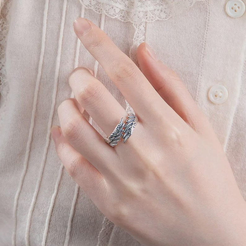 Puede incluir: Un anillo plateado con un dise&ntilde;o de plumas. El anillo envuelve el dedo, con los detalles de las plumas visibles en la parte superior. El anillo se lleva en el dedo de una persona, con un fondo suave de tonos neutros.