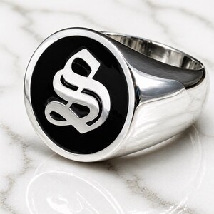 May include: A silver signet ring with a black enamel face. The face features a stylized, white letter "S". The ring is highly polished and reflects the light. The ring is likely made of sterling silver.