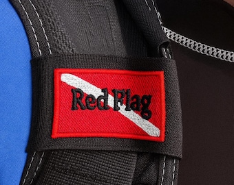 May include: A rectangular embroidered patch featuring a red background with a diagonal white stripe and the black text Red Flag. The patch is attached to a black fabric strap with white stitching, positioned next to a blue and black garment.