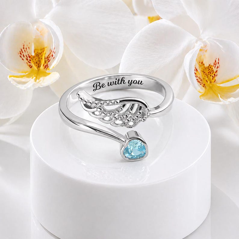 Puede incluir: Un anillo plateado con dise&ntilde;o de ala y una piedra preciosa azul. El anillo est&aacute; grabado con las palabras "Be with you". El anillo se muestra sobre una superficie blanca con flores blancas en el fondo.