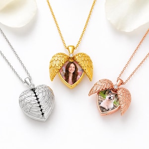 May include: Three heart-shaped locket necklaces with wing designs. One is silver, one is gold, and one is rose gold. The gold and rose gold lockets are open, revealing photos. Each necklace has a delicate chain.