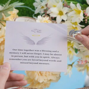 Puede incluir: Un anillo plateado con dise&ntilde;o de ala se sostiene frente a una tarjeta con texto. La tarjeta dice "Hope Rings" e incluye un mensaje sobre el amor y el recuerdo. El fondo presenta flores blancas y amarillas.