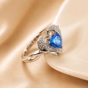 May include: A silver ring with a heart-shaped blue gemstone at the center. The ring features a heart-shaped design with small, clear stones and the inscription "Always in my heart".