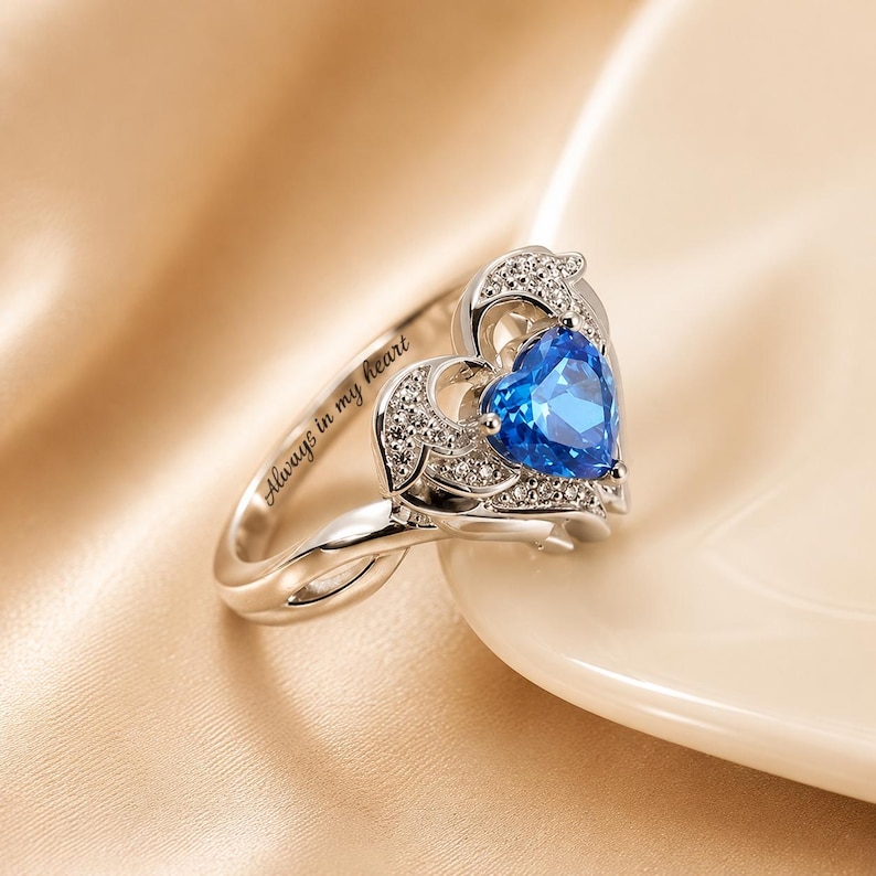 May include: A silver ring with a heart-shaped blue gemstone at the center. The ring features a heart-shaped design with small, clear stones and the inscription "Always in my heart".