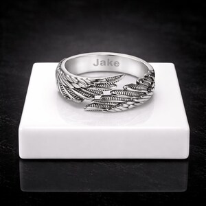 Puede incluir: Un anillo plateado con dise&ntilde;o de alas. El anillo est&aacute; grabado con el nombre "Jake". Las alas envuelven la banda, creando un aspecto &uacute;nico y detallado. El anillo se muestra sobre una superficie blanca.