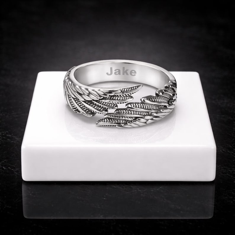 Puede incluir: Un anillo plateado con dise&ntilde;o de alas. El anillo est&aacute; grabado con el nombre "Jake". Las alas envuelven la banda, creando un aspecto &uacute;nico y detallado. El anillo se muestra sobre una superficie blanca.