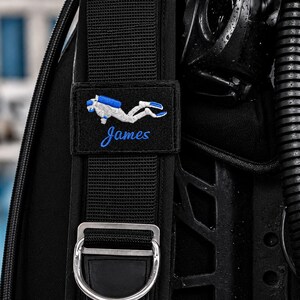 Può includere: Una cinghia nera per giubbotto ad assetto variabile da sub con una toppa rettangolare che presenta un'icona ricamata di un subacqueo bianco e blu e il nome James in corsivo blu, posizionata sopra un punto di attacco a D in metallo argentato.