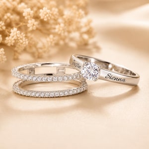 May include: A silver-toned ring set featuring a solitaire ring with a round clear stone and the engraved names Justin and Sienna on the band, accompanied by a matching eternity band encrusted with small clear stones along the entire circumference.