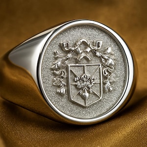 May include: A silver signet ring with a detailed crest design. The ring features a round face with a textured background and a raised emblem of a shield, floral elements, and other heraldic symbols. The ring is set against a gold background.