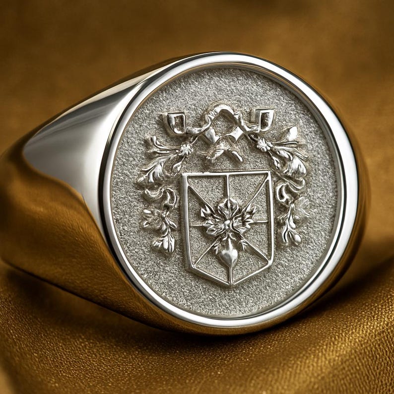 May include: A silver signet ring featuring a circular face with a textured background. The center displays an engraved shield emblem with a floral motif, surrounded by ornate scrollwork detailing that extends above the shield.