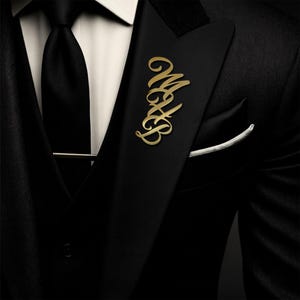 Op de afbeelding: Een goudkleurige reversspeld met de initialen "MAB" in een elegant lettertype is bevestigd aan een zwart colbert. De jas heeft een zwarte stropdas, een wit overhemd en een pochet. De speld voegt een vleugje verfijning toe.