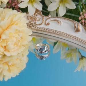 Puede incluir: Un anillo plateado con dise&ntilde;o de plumas, sobre una superficie azul reflectante. El anillo tiene grabado el nombre "Andre". El fondo incluye flores blancas y amarillas y un marco decorativo blanco y dorado.
