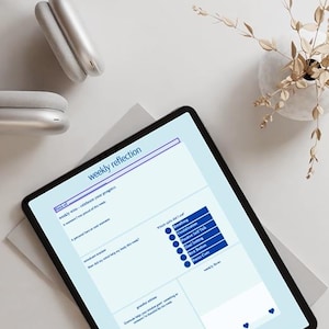 Puede incluir: Una tableta digital que muestra una plantilla de "Reflexión Semanal" con secciones para la atención plena, las habilidades y la gratitud. También se ven unos auriculares blancos y un pequeño jarrón con plantas secas.