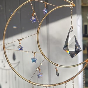 May include: A celestial-themed suncatcher featuring a gold-colored crescent moon frame. The design includes dangling star-shaped and teardrop-shaped crystals, along with a large, faceted crystal pendant. The suncatcher is suspended by a delicate chain.