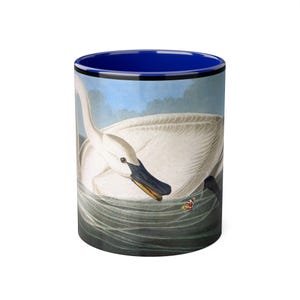 Puede incluir: Taza de cerámica blanca con una ilustración detallada de un cisne en el agua. El cisne tiene un pico negro y está sobre un fondo de cielo azul y agua. El interior de la taza es azul.