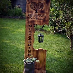 Könnte beinhalten: Ein rustikales Holzschild mit den Worten "Willkommen bei Familie" und "Mustermann" eingraviert. Das Schild zeigt ein herzförmiges Baumdesign und eine kleine schwarze Laterne. Am Fuß des Schildes steht eine Topfpflanze.