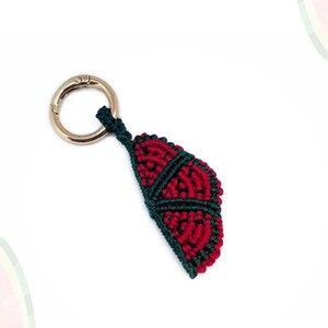 Puede incluir: Un llavero de macramé con forma de rebanada de sandía con un cierre dorado. La rebanada de sandía es roja con detalles en verde oscuro, y el texto "LUMETHRE WATERMELON COLLECTION" está en la parte superior.