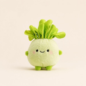 May include: A plush toy shaped like a round, light green vegetable with a cluster of upright, vibrant green leaves on top. The toy features a stitched black smile, two small black eyes, and soft pink blush marks on its cheeks, with tiny green limbs.