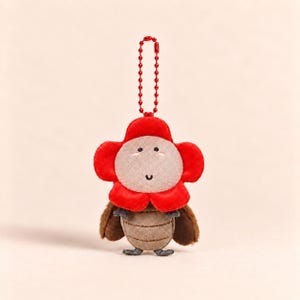 Op de afbeelding: Een pluche sleutelhanger met een figuurtje met een lichtbeige rond gezicht, een felrode bloemvormige kraag en een bruin gesegmenteerd lichaam. Het figuurtje heeft geborduurde ogen en een mond, met een rode metalen kogelketting aan de bovenzijde.