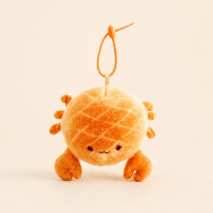 Puede incluir: Un adorno de peluche con forma de cangrejo naranja, textura suave y un patrón de cuadrícula en su cuerpo redondo. El cangrejo tiene dos ojos oscuros pequeños, una boca diminuta, tres patas pequeñas a cada lado y dos pinzas delanteras.