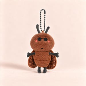 May include: A brown plush insect keychain featuring a fuzzy round head with black circular sunglasses, small pink cheeks, and two antennae. The body has a segmented texture, dark gray limbs, and brown wings with a repeating gold geometric pattern.