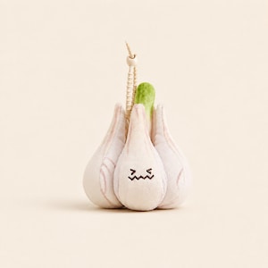 Puede incluir: Un juguete de peluche con forma de cabeza de ajo con un exterior de color blanco roto con sutiles tonos rosados, un pequeño brote verde en la parte superior y una cara bordada en marrón con una boca en zigzag y ojos cerrados. Un cordón beige está unido arriba.