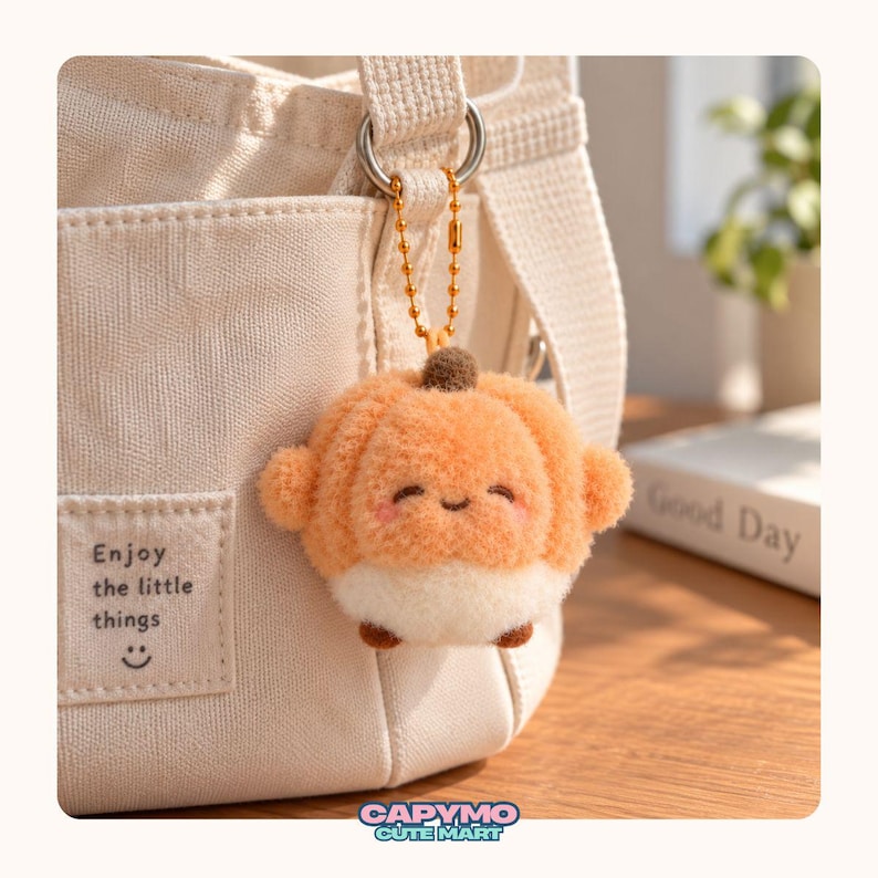 Puede incluir: Un llavero de peluche con forma de calabaza naranja y blanca con un peque&ntilde;o tallo marr&oacute;n, sujeto a un bolso de lona color crema mediante una cadena de bolas dorada. El bolso tiene un parche rectangular con el texto Enjoy the little things.