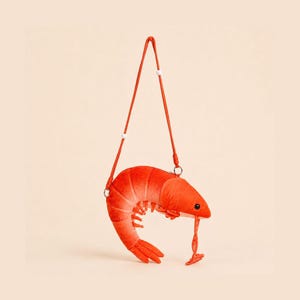 May include: A plush shrimp-shaped crossbody bag in a vibrant orange colour. The bag features a long, thin matching orange strap with small white beads, a curved body design, and a small black eye detail on the side of the shrimp's head.