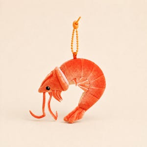 Puede incluir: Un llavero de peluche con forma de camarón que presenta un cuerpo de tela naranja vibrante con una textura suave. El camarón tiene una cola curvada, ojos negros pequeños y antenas largas. Una cadena de bolas naranja está unida en la parte superior.