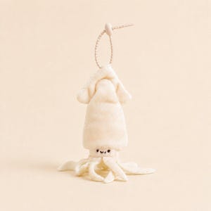 May include: A cream-colored plush squid toy with a soft, velvety texture. The squid features a simple embroidered face with two small black eyes and a curved mouth. A thin, matching cream-colored beaded cord is attached to the top of the head.