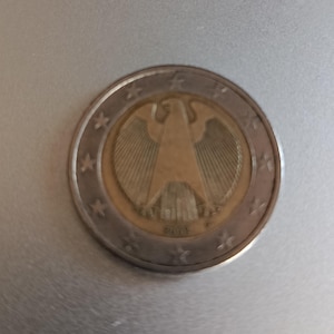 May include: A two-euro coin with a gold-coloured eagle emblem in a gold-coloured centre. The outer ring is silver with twelve stars. The coin's design includes the year 2005. This coin is a form of currency.