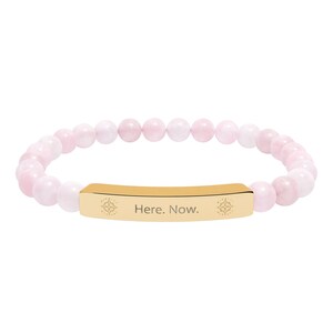 Op de afbeelding: Een elastische armband met lichtroze, bolvormige kralen en een centrale goudkleurige metalen staaf. De staaf is gegraveerd met de tekst "Here. Now." en is aan weerszijden van de inscriptie voorzien van een klein decoratief bloemembleem.