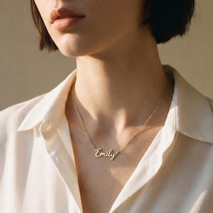 May include: A gold-toned metal necklace featuring a delicate chain and a central nameplate pendant with the name Emily written in a cursive script font. The necklace rests against the neck, showcasing a polished finish and a simple link chain design.