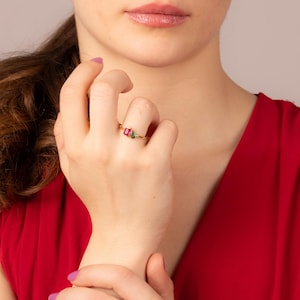 May include: A gold ring with a rectangular red gemstone and a teardrop-shaped green gemstone. The ring is displayed on a person's finger. The background is a solid red colour.