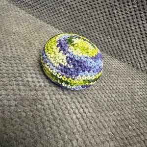 May include: A crocheted ball in shades of green, blue, and purple. The handmade ball is round and has a textured surface, with the different colors creating a striped pattern. The ball is sitting on a gray surface.