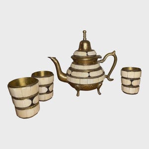 May include: A brass and bone inlay tea set, including a teapot and three matching glasses. The teapot has a curved spout and handle, with a decorative lid. The glasses and teapot are decorated with white bone inlay and brass accents.