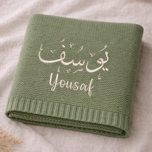 May include: A folded, sage green knitted blanket with the name "Yousaf" embroidered in white, both in Arabic script and English lettering. The blanket has a ribbed border and is displayed on a neutral-colored fabric.