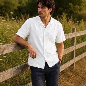 May include: A white short-sleeved button-down shirt with a camp collar and a straight hemline, paired with dark navy blue trousers. The shirt features a visible button placket down the center and a textured fabric finish.