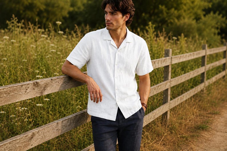 May include: A white short-sleeved button-down shirt with a camp collar and a straight hemline, paired with dark navy blue trousers. The shirt features a visible button placket down the center and a textured fabric finish.
