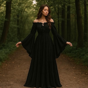Puede incluir: Un vestido largo negro con hombros descubiertos y mangas fluidas. El vestido tiene un corpi&ntilde;o ajustado y una falda amplia. La modelo est&aacute; de pie en un camino en un bosque, con &aacute;rboles en el fondo. El vestido est&aacute; hecho de una tela suave y fluida.