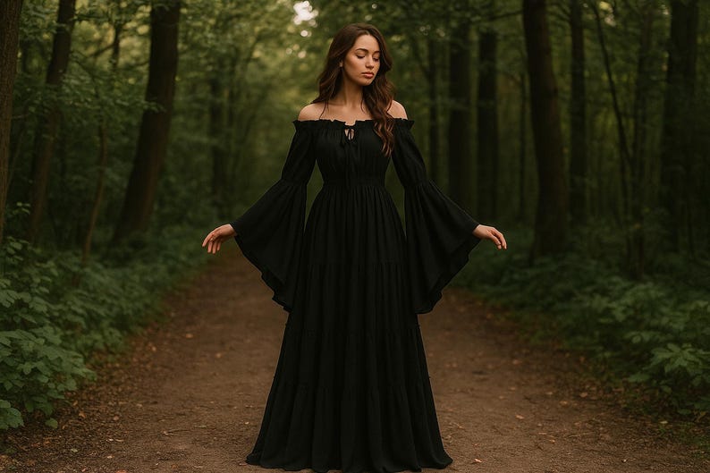 Puede incluir: Un vestido largo negro con hombros descubiertos y mangas fluidas. El vestido tiene un corpi&ntilde;o ajustado y una falda amplia. La modelo est&aacute; de pie en un camino en un bosque, con &aacute;rboles en el fondo. El vestido est&aacute; hecho de una tela suave y fluida.