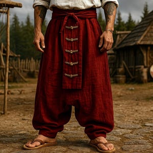 May include: Burgundy-colored linen pants with a wide waistband and decorative gold-colored clasps. The pants have a loose, baggy fit and are worn with brown leather sandals. The background features rustic wooden structures and a forest.