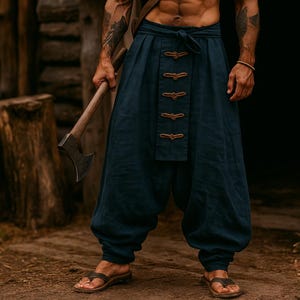 May include: Navy blue linen pants with decorative brown closures. The pants are loose-fitting and gathered at the ankles. The person is wearing brown leather sandals and holding an axe. The background features a wooden structure and tree stumps.