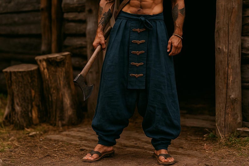May include: Navy blue linen pants with decorative brown closures. The pants are loose-fitting and gathered at the ankles. The person is wearing brown leather sandals and holding an axe. The background features a wooden structure and tree stumps.