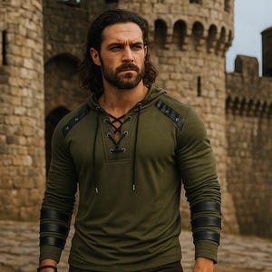 May include: An olive green hooded long-sleeve shirt with black leather accents on the shoulders and sleeves. The shirt features a lace-up neckline and is worn by a person standing in front of a stone castle.